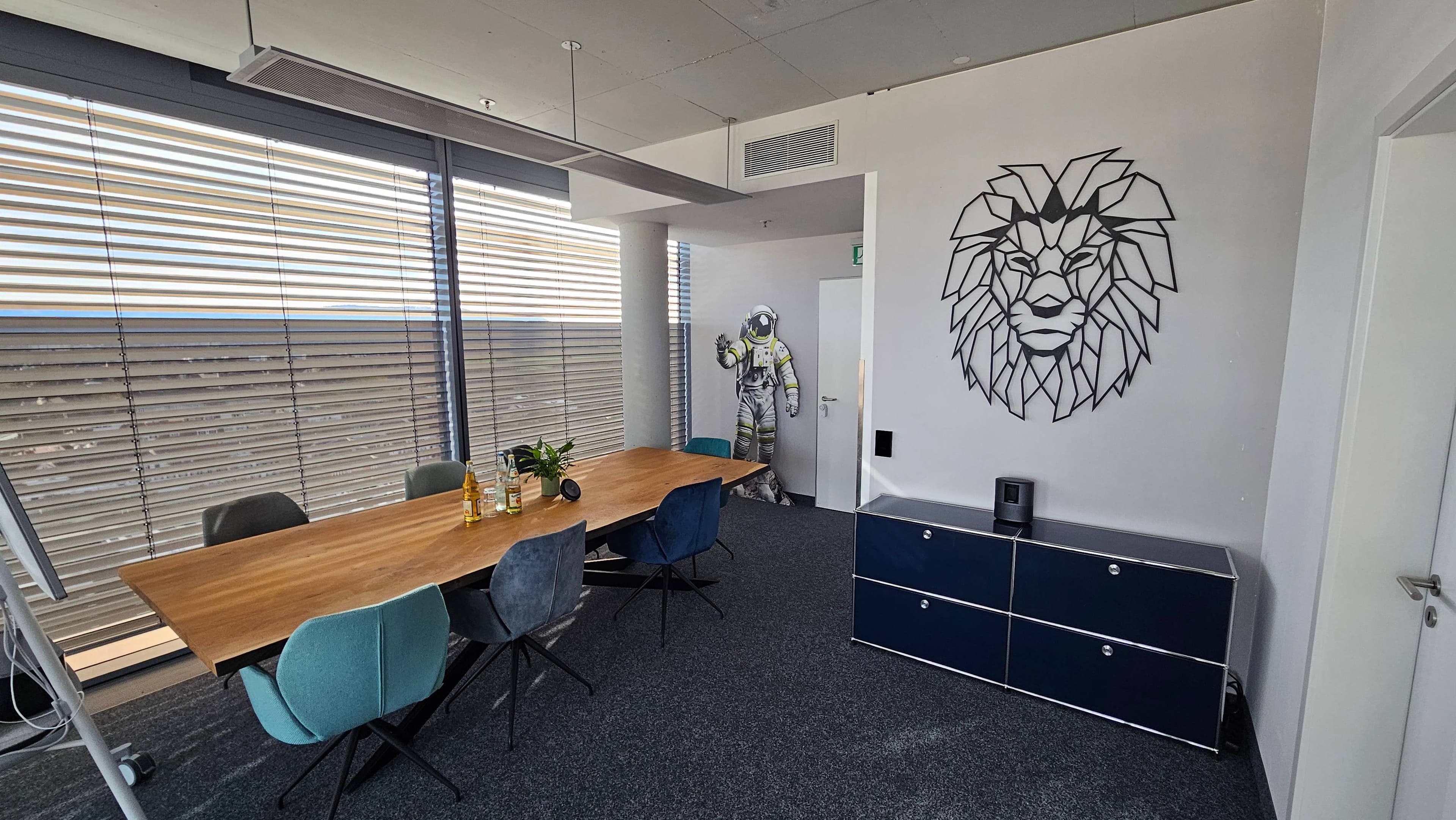 Besprechungsraum im neuen NMS-Büro mit langem Holztisch, blauen und türkisen Stühlen und einem Löwenkopf aus Metall an der Wand