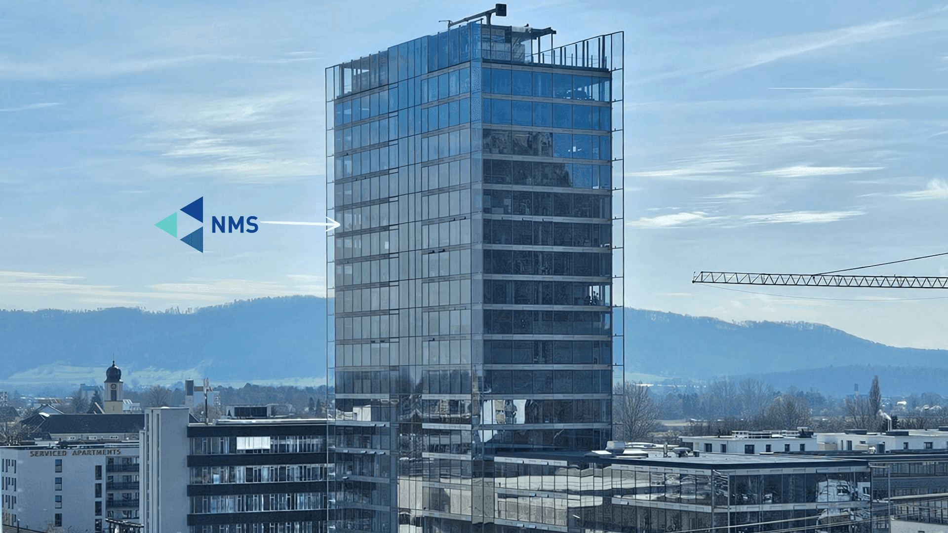Der Hegau Tower, ein verglastes Hochhaus, mit dem NMS-Logo und einem Pfeil auf das Bürogeschoss; im Hintergrund ein Kirchturm und Hügel