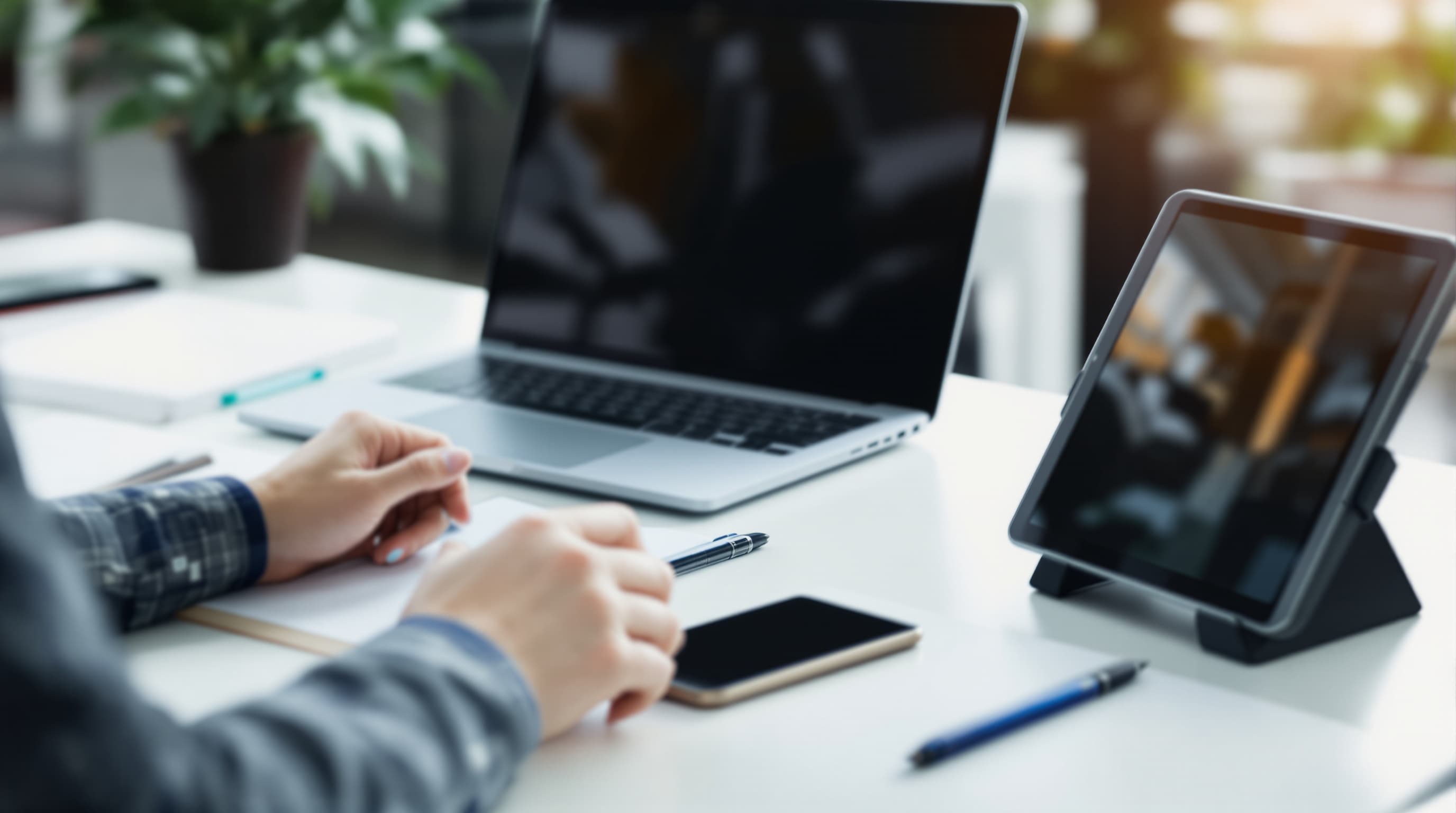 Arbeitsplatz mit Laptop, Tablet und Smartphone, im Vordergrund Hände mit Stift und Notizbuch